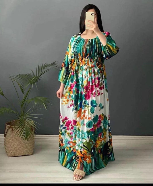 Woman wearing a colorful floral dress taking a mirror selfie against a gray wall with a plant on the left.